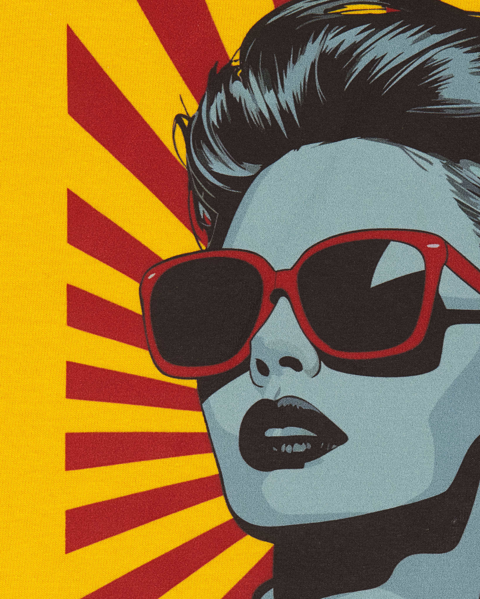 Zbliżenie na miękki nadruk pop art na żółtym t-shircie damskim. Grafika przedstawia twarz kobiety w czerwonych okularach przeciwsłonecznych na tle czerwono-żółtych promieni. Widoczna struktura materiału bawełnianego.