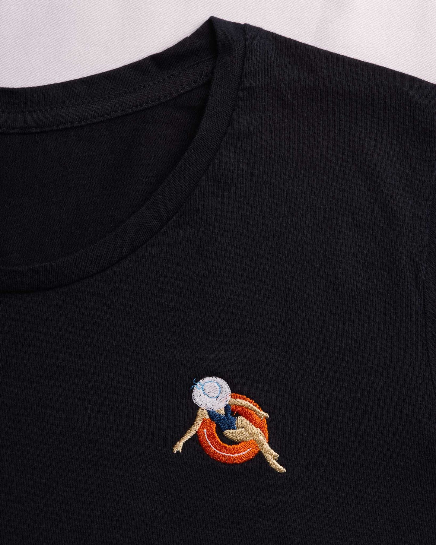 Zbliżenie na granatowy t-shirt damski premium z małym, kolorowym haftem komputerowym na piersi, przedstawiającym kobietę w kapeluszu wypoczywającą na pomarańczowym kole ratunkowym.