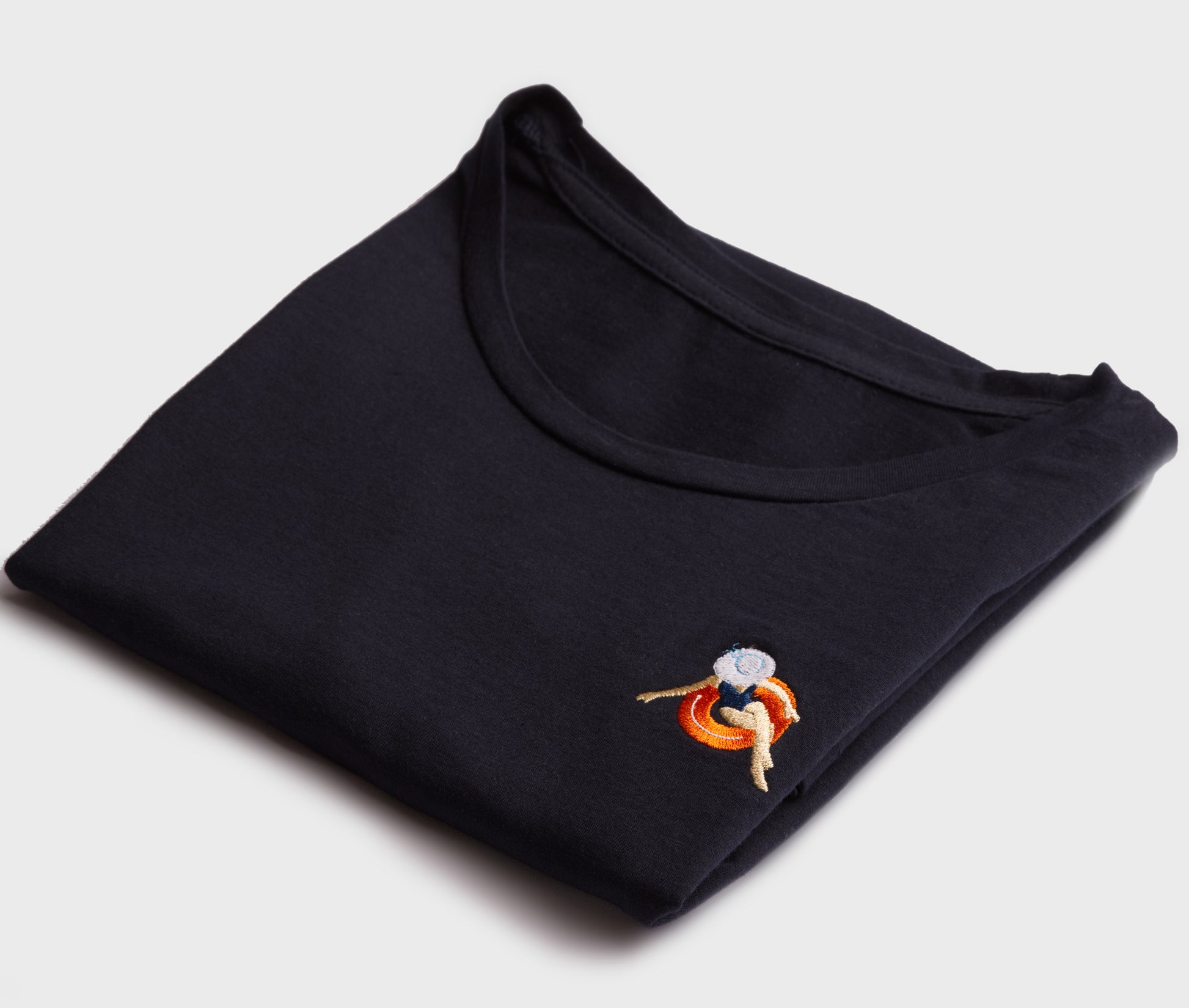 Starannie złożony, granatowy t-shirt damski premium, prezentujący mały, kolorowy haft komputerowy na piersi z motywem kobiety wypoczywającej na pomarańczowym kole ratunkowym.